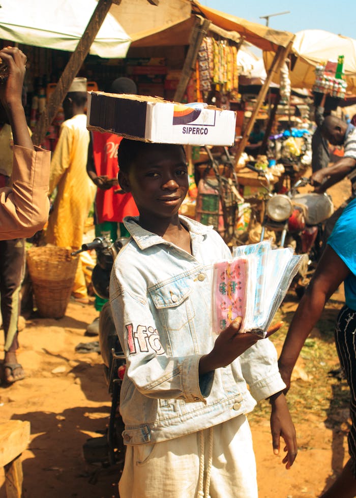 gallery-03 A young vendor selling goods with a box on his head in a vibrant outdoor market.